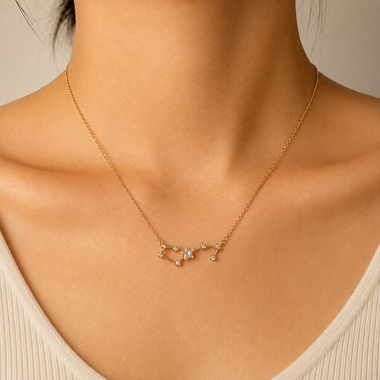 A close-up of a model's neck and upper chest wearing a delicate gold chain necklace with a small constellation pendant set with clear gemstones, paired with a cream-colored ribbed top.
