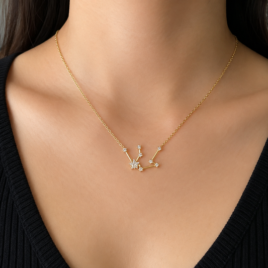 A gold necklace with a constellation pendant featuring small clear gemstones, worn on a model's neck over a black V-neck top.
