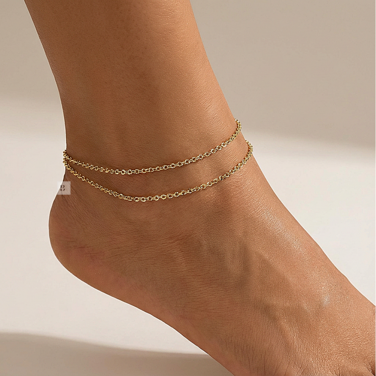 Gold anklets on a person's ankle against a neutral background