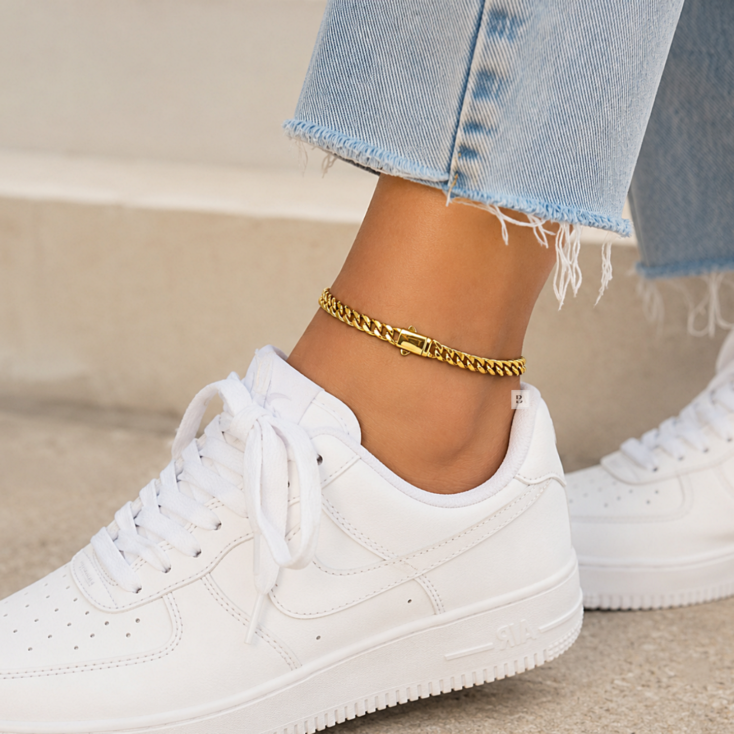 A close-up of a person's ankle wearing a gold chain anklet, paired with light blue frayed-hem jeans and white low-top sneakers.