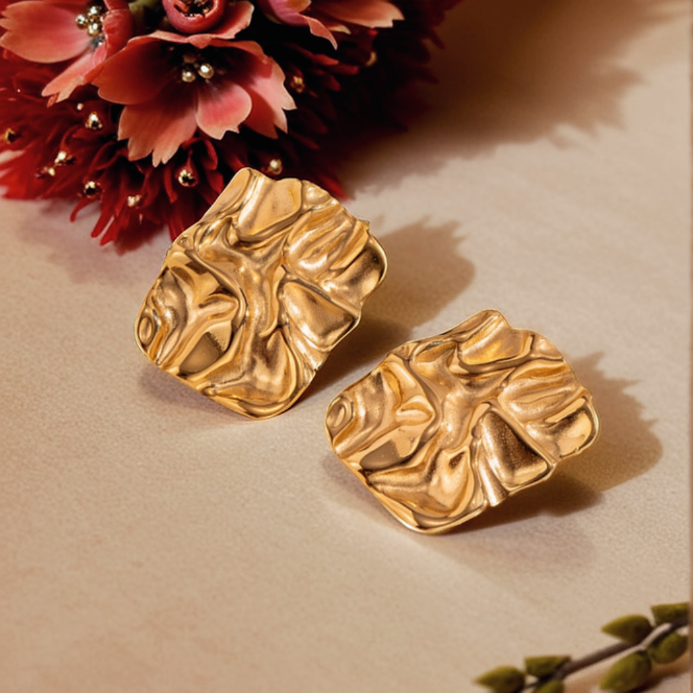 Gold leaf-shaped earrings on a beige surface with pink flowers in the background