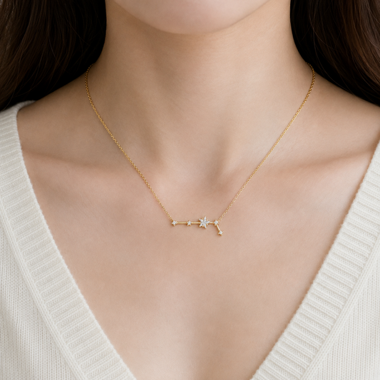 A delicate gold necklace with a small celestial or star-like pendant, worn by a model in a close-up shot. The model is wearing a cream-colored ribbed knit top.