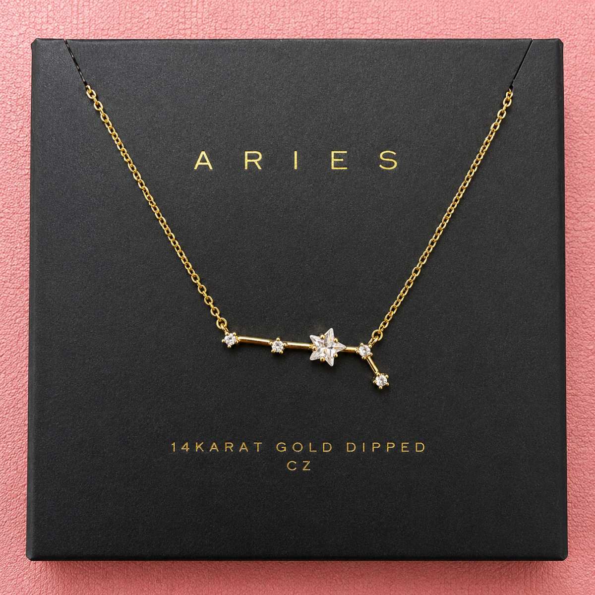 A gold necklace with a constellation pendant displayed on a black gift box. The necklace features a star and constellation design with clear gemstones, and the box is printed with the name 'ARIES' and '14KARAT GOLD DIPPED CZ'.