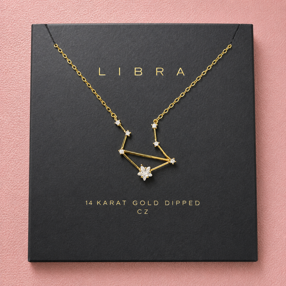 A gold-dipped necklace with a constellation pendant and star accents, displayed on a black square gift box printed with the name LIBRA and product text, set against a pink background.
