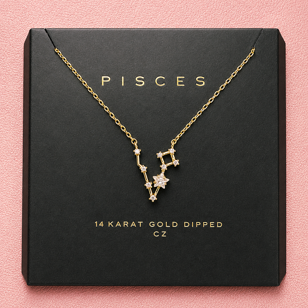 A gold necklace with a Pisces constellation pendant displayed on a black presentation box against a pink textured background. The box is printed with the brand name 'PISCES' and text reading '14 KARAT GOLD DIPPED CZ'.