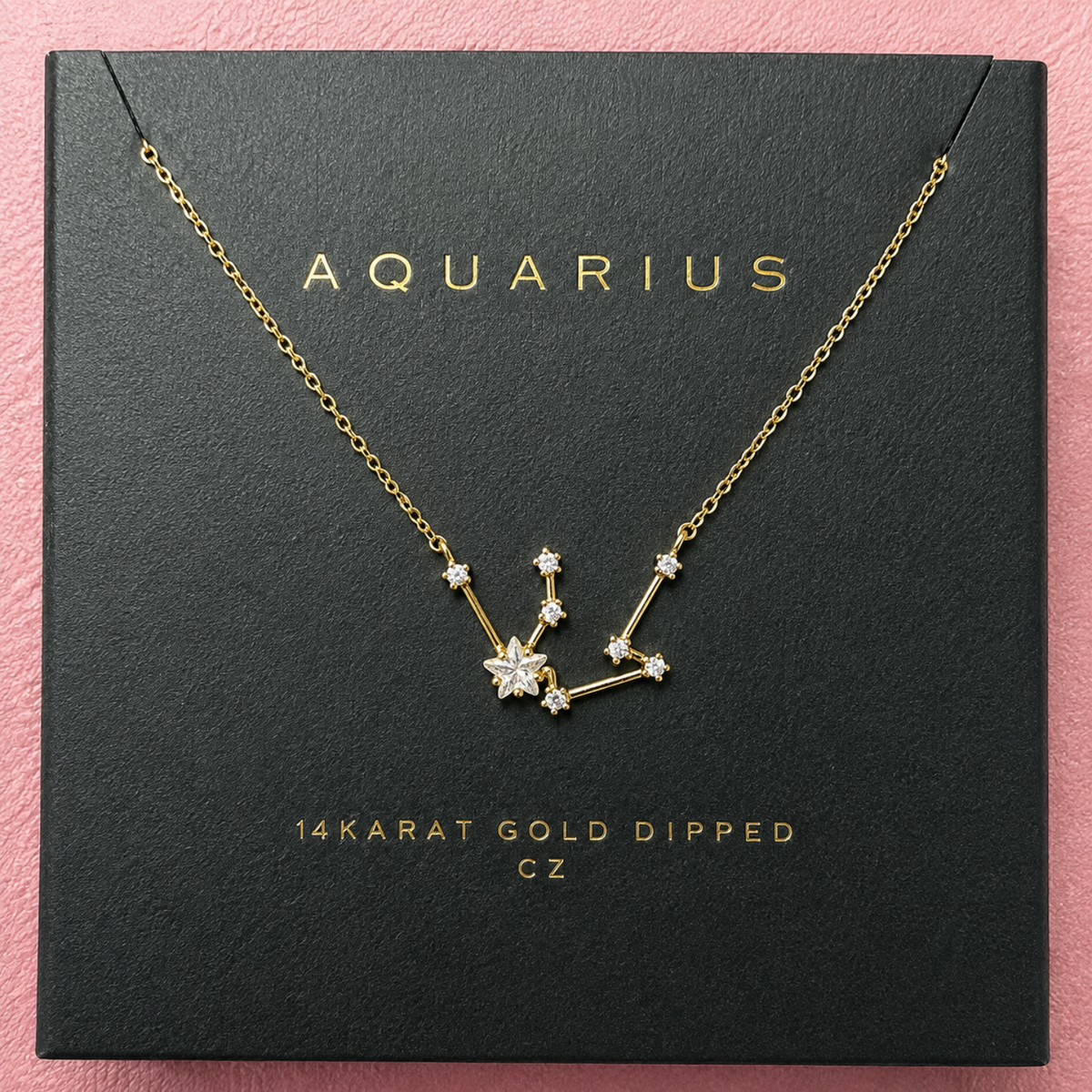 A gold Aquarius constellation necklace with clear gemstones, presented on a black textured square box with gold lettering, set against a pink background.