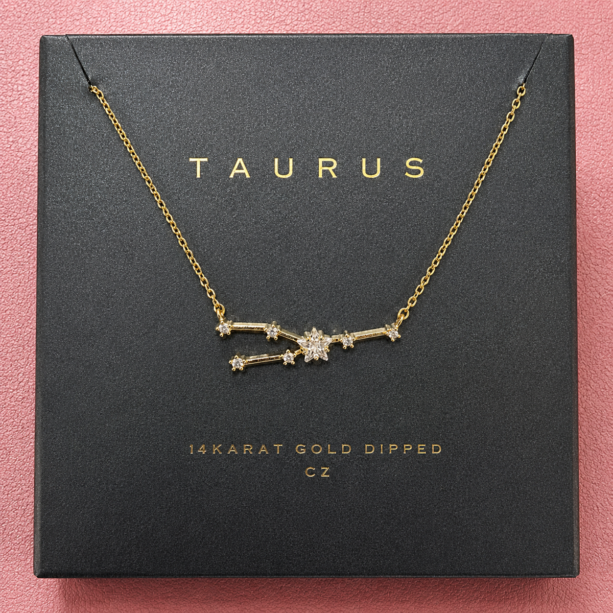 A gold necklace with a Taurus constellation pendant, displayed on its black presentation box which features the brand name 'TAURUS' in gold lettering, set against a textured pink background.