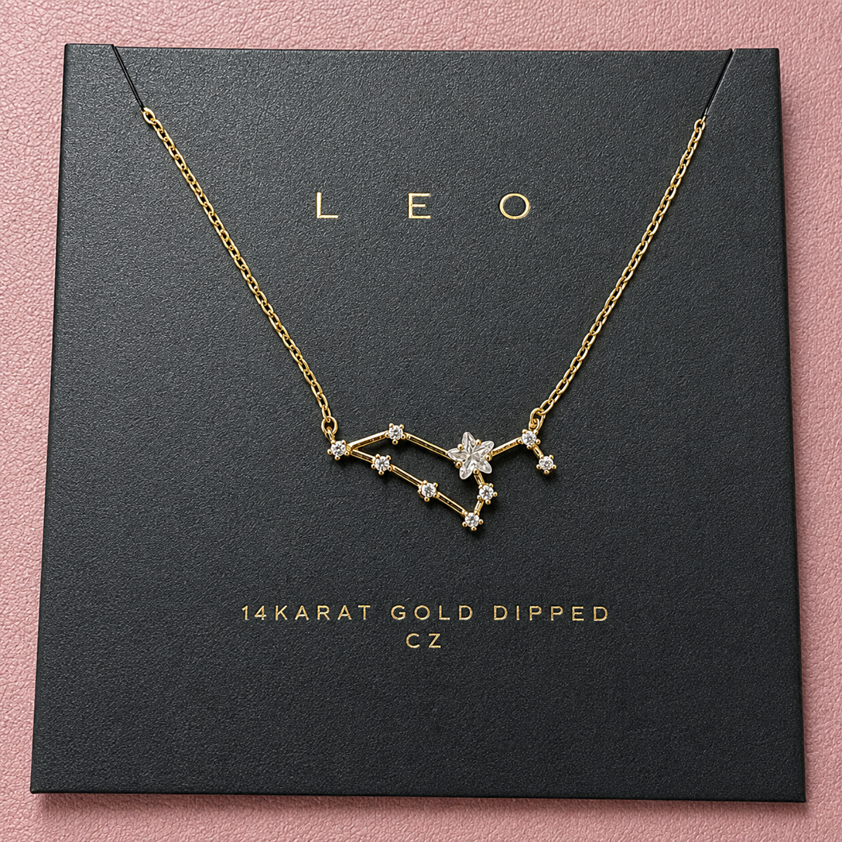 A gold-dipped necklace with a Leo constellation pendant, displayed on a black gift box with gold lettering against a pink background.