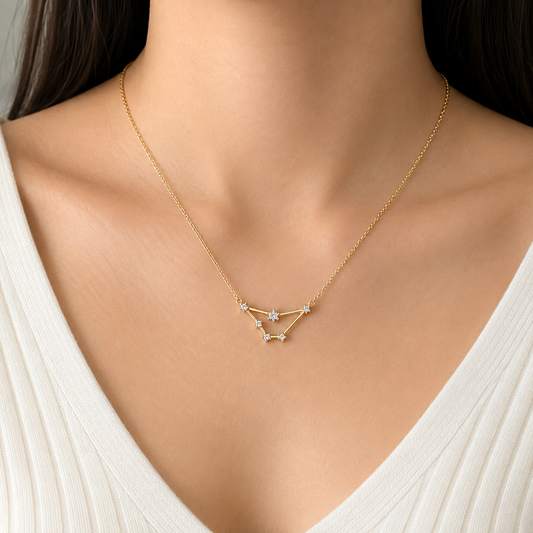 A close-up of a model wearing a delicate gold necklace with a geometric constellation pendant. The necklace features a fine gold chain and a pendant composed of connected gold lines and small star-shaped elements, each accented with a clear gemstone.