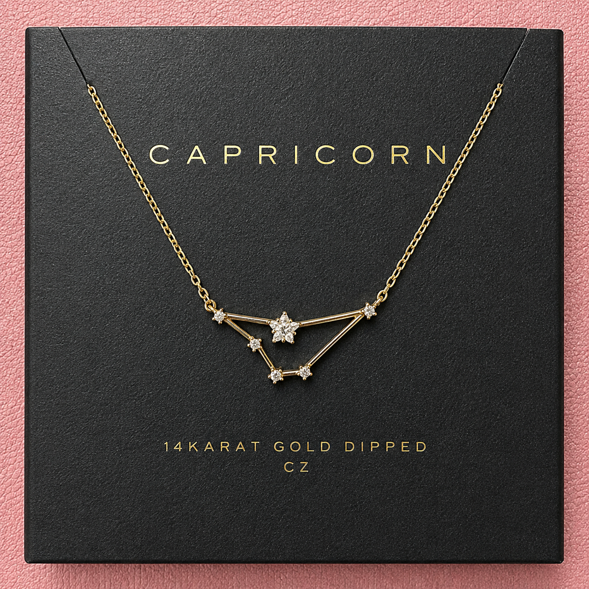 A gold-dipped necklace with a Capricorn constellation pendant, displayed on its black branded packaging box against a pink background.
