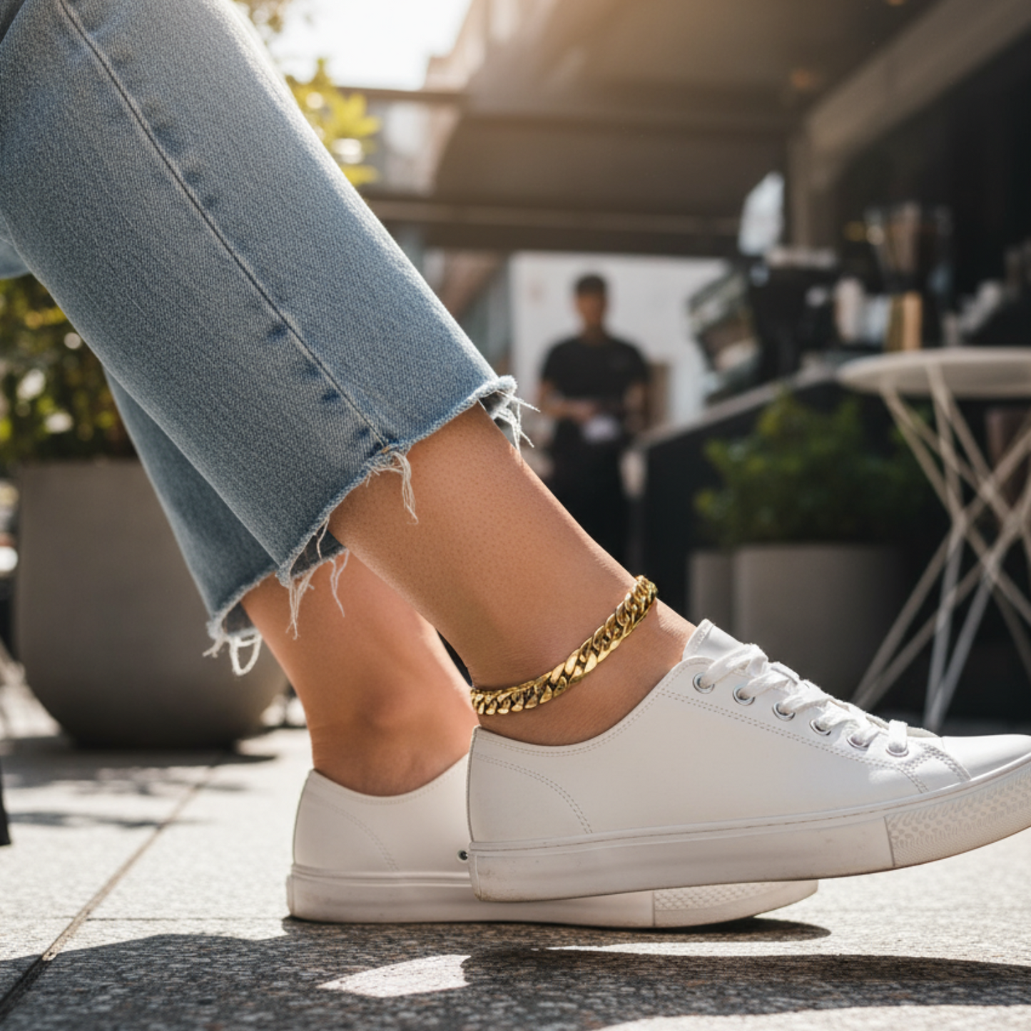 Person wearing white sneakers and a gold anklet, standing on a sidewalk with a blurred background.