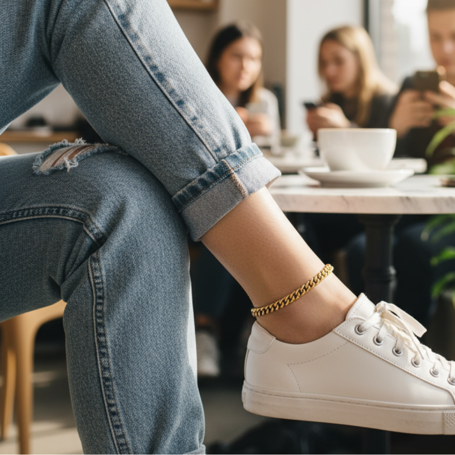 Person wearing white sneakers and ripped jeans sitting in a cafe with blurred people in the background.