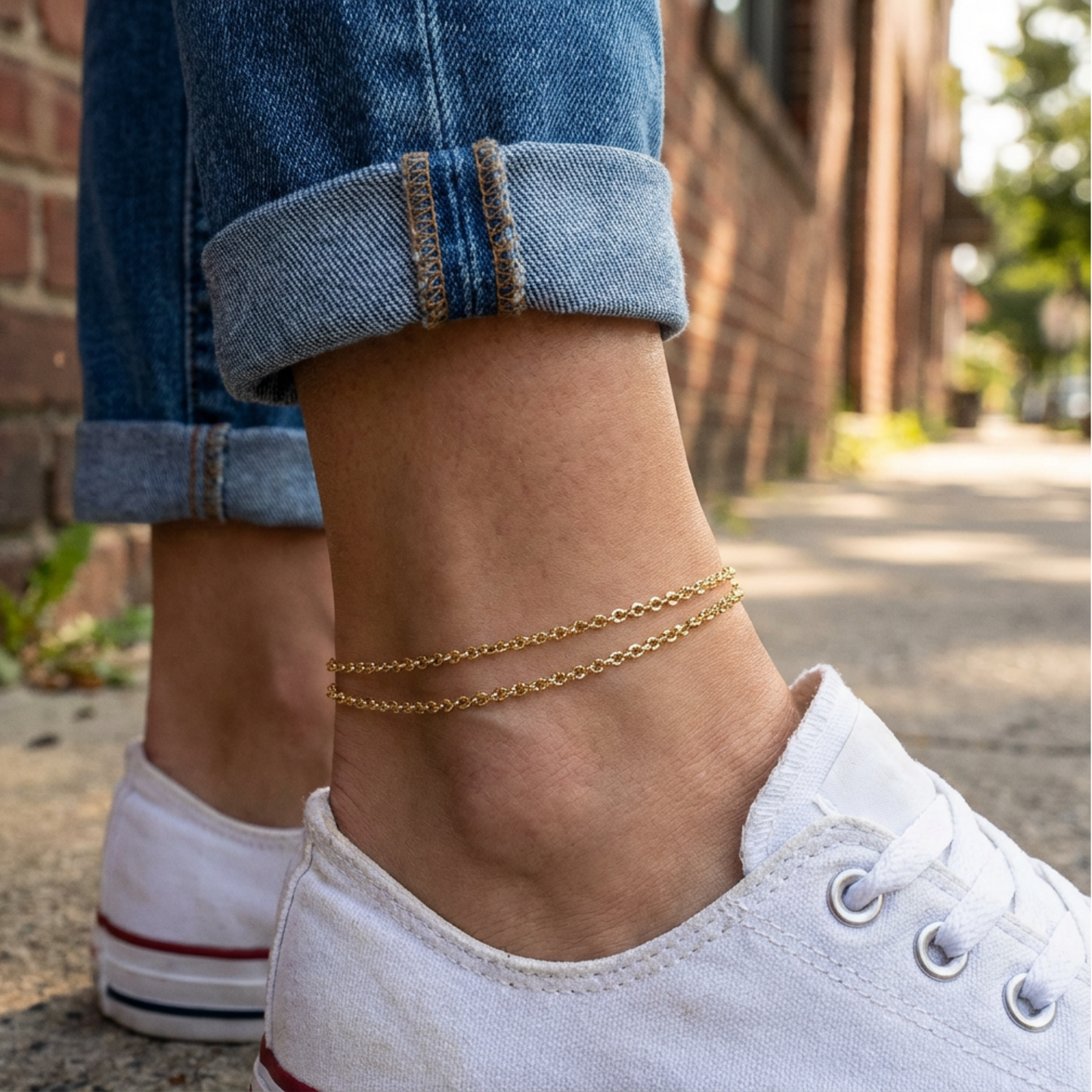 Person wearing gold anklets on a sidewalk with rolled-up jeans and white sneakers.