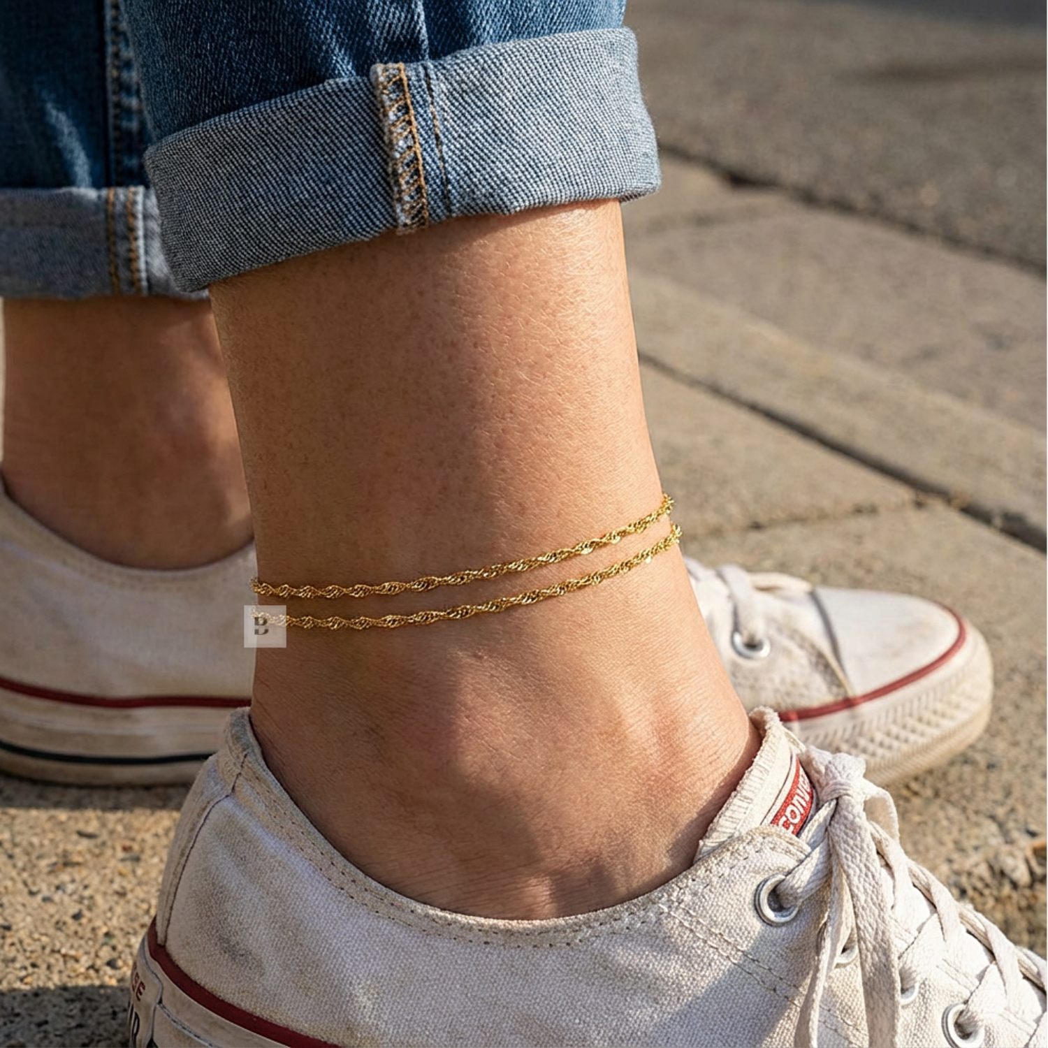 Close-up of a person's ankle with gold anklets wearing white sneakers on a sidewalk.
