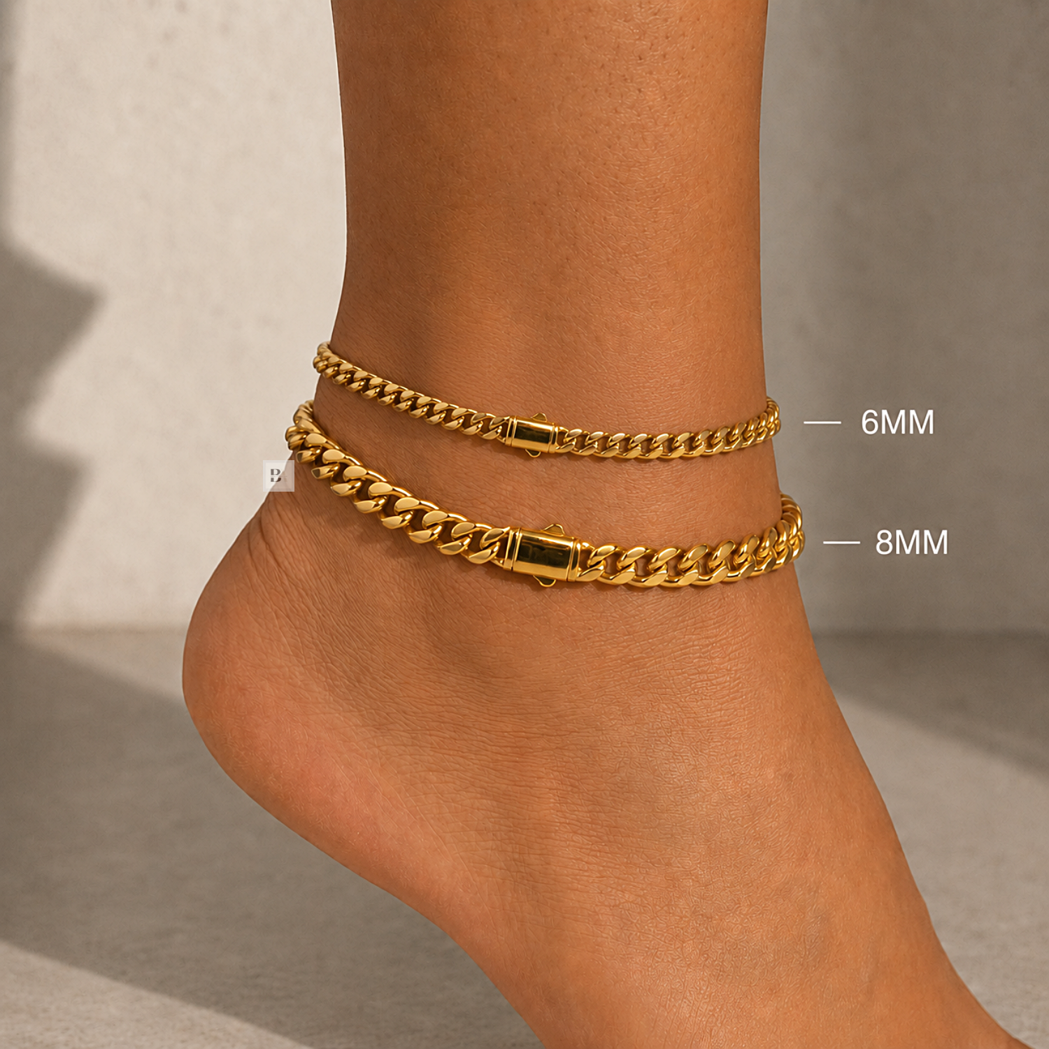 A close-up of a person's ankle and foot wearing two gold chain anklets, one labeled 6MM and one labeled 8MM, against a neutral light gray background.