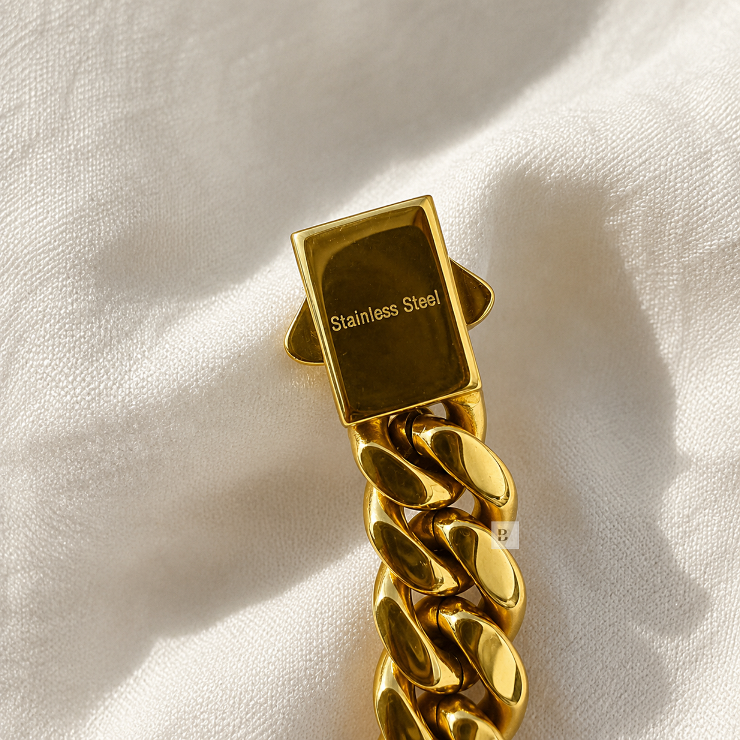 A close-up of a gold-toned stainless steel chain link bracelet clasp resting on a textured white fabric background.