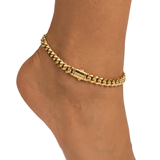 A close-up of a gold-toned Cuban link chain anklet worn on a person's ankle, shown against a plain white background.