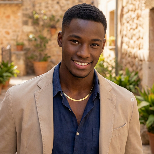 Man wearing a Gold Herringbone Necklace beige blazer over a blue shirt in an outdoor setting with plants and stone wall.