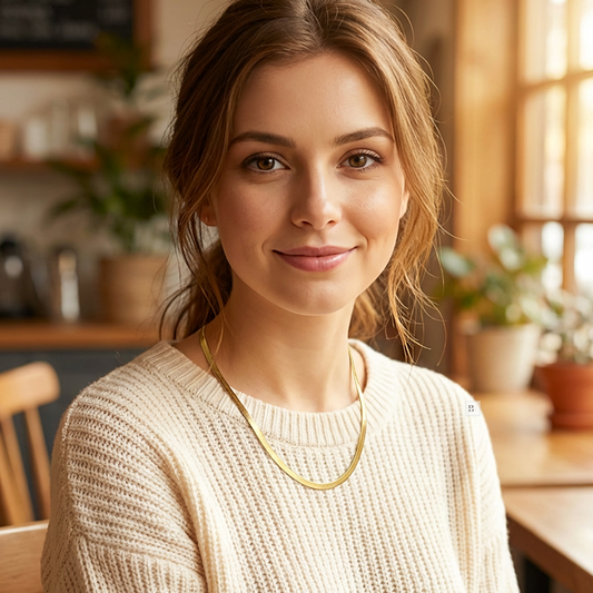 Woman wearing a beige sweater with a gold necklace in a warm, indoor setting.